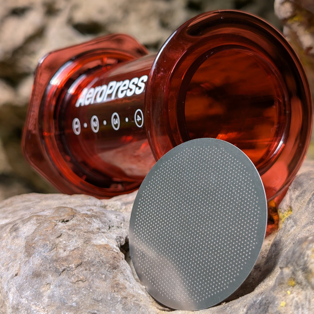 Das Bild zeigt eine Nahaufnahme eines Metallfilters für die AeroPress - Kaffeezubereiter. Im Hintergrund ist eine rote AeroPress mit weißer Aufschrift zu erkennen. Die Umgebung besteht aus grau braunen Felsformationen.