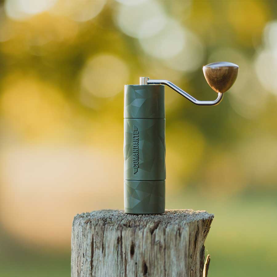 Das Bild zeigt eine Comandante Trailmaster X25 Forest zusammengebaut, stehend auf einem Baumstumpf. Der grüne und Hellgrüne Hintergrund ist unscharf und lässt den Fokus auf der Handmühle ruhen.