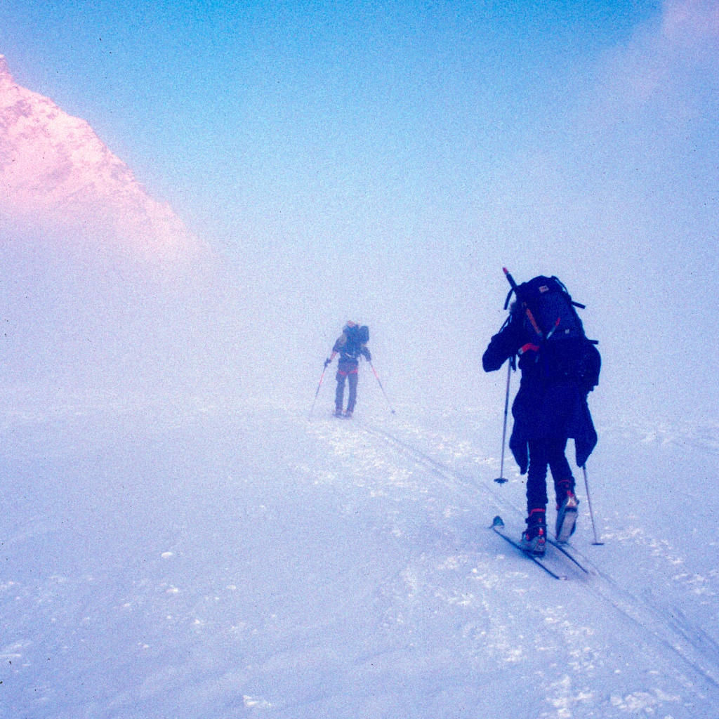 Das Bild zeigt zwei Bergsteiger auf Skiern. Der Untergrund ist weiß und Nebel verhindert die Sicht. Links oben kann man leicht einen, von der Sonne angestrahlten Bergrücken sehen.
