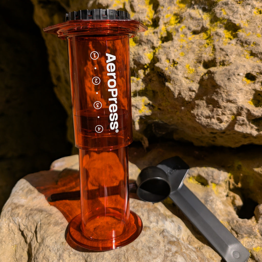 Das Bild zeigt eine rötlich schimmernde AeroPress Kaffeezubereiter welcher strahlend auf einer hellbraunen Felswand steht. Im Hintergrund liegt ein schwarzer Löffel und ein schwarzer Rührstab.