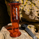Das Bild zeigt eine rötlich schimmernde AeroPress Kaffeezubereiter welcher strahlend auf einer hellbraunen Felswand steht. Im Hintergrund liegt ein schwarzer Löffel und ein schwarzer Rührstab.