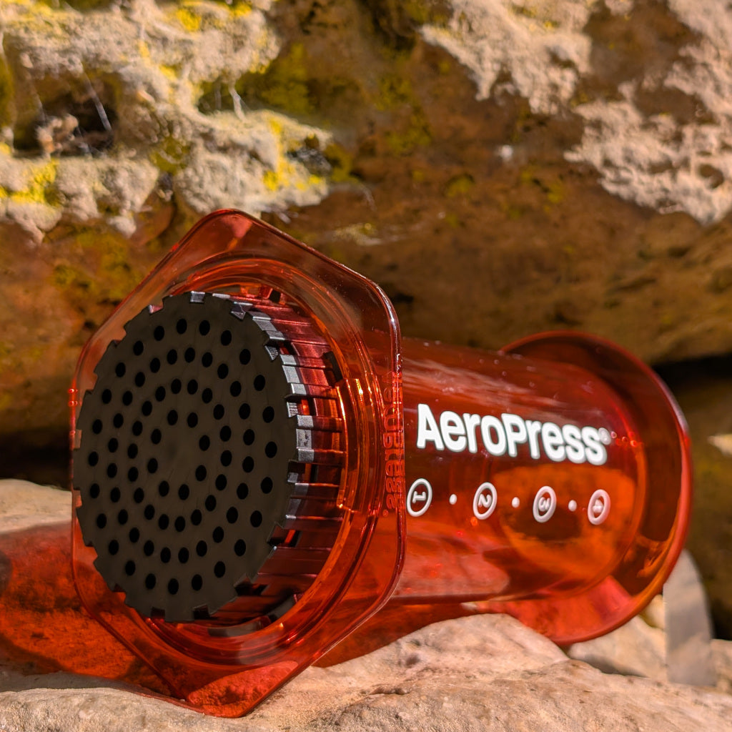 Das Bild zeigt eine Nahaufnahme einer AeroPress red clear mit der weißen Aufschrift AeroPress. Der Kaffeebereiter liegt auf einer Felsoberfläche und im Hintergrund sind hellbraune Felsformationen zu erkennen. 