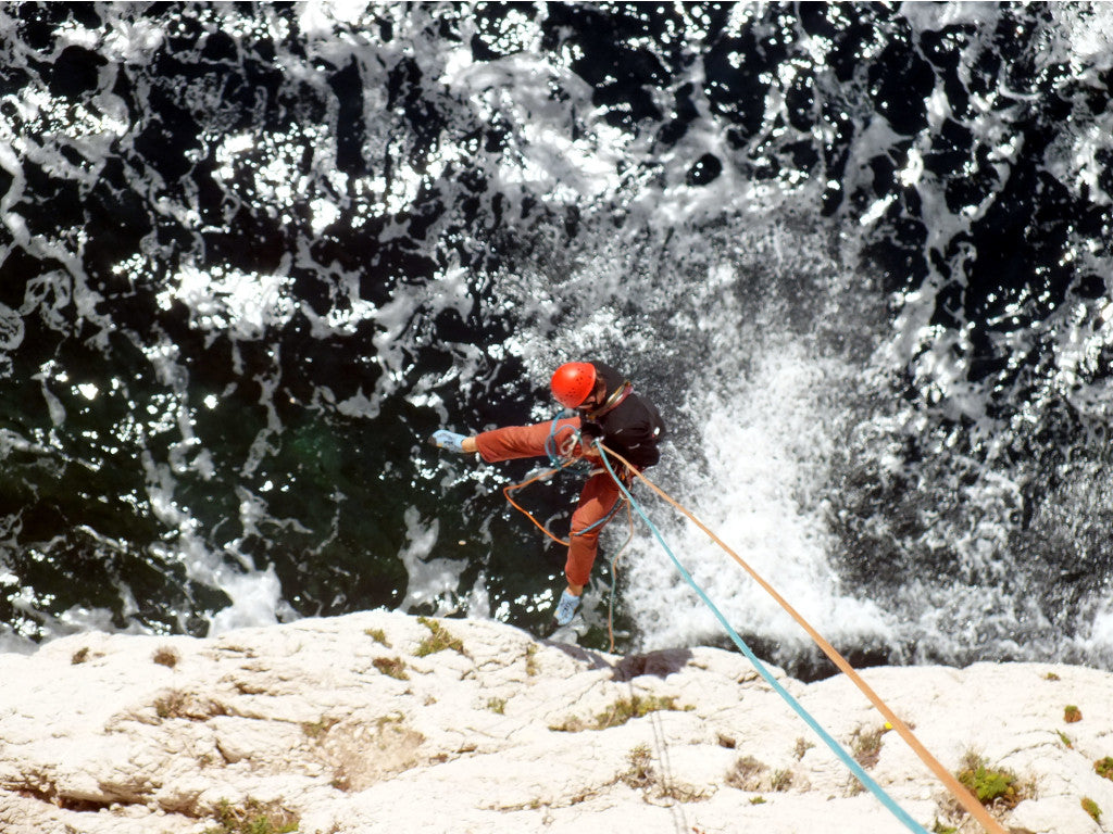 Das Bild zeigt Felsformationen und spritzende Meeresgischt. Im Vordergrund ist ein Kletterer zu sehen der sich an einem orangenem und blauen Seil Richtung Wasser abseilt. 
