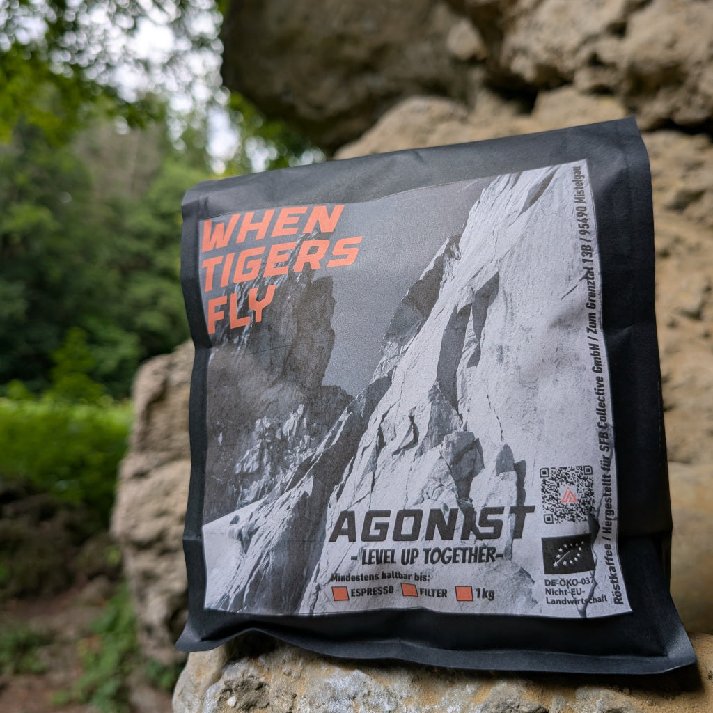 Das Bild zeigt eine schwarze Kaffeeverpackung mit dem grau, orangenen Etikett von WHEN TIGERS FLY auf einem Felsvorsprung stehend. Im Hintergrund sind unscharf Felsen und Wald zu erkennen. 