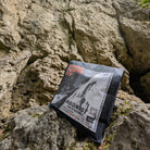 Das Bild zeigt eine schwarze Kaffeeverpackung mit dem grau, orangenen Etikett von WHEN TIGERS FLY. Die Verpackung steht auf einem Felsvorsprung und im Hintergrund ist eine Felswand zu sehen welche mit Rissen und Löchern durchzogen ist. 