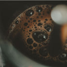 Das Bild zeigt eine Nahaufnaheme von Kaffeeschaum auf Kaffee in einer Tasse. Der braune Schaum bildet Blasen in welchen sich das Licht spiegeln. 