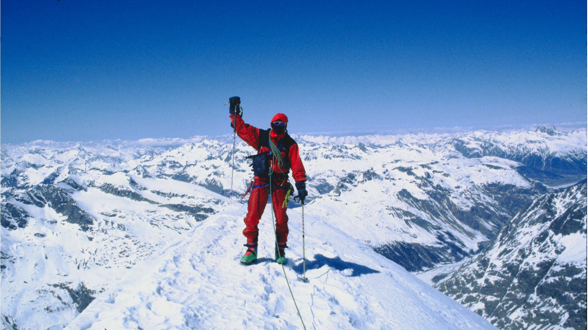 Das Bild zeigt einen Bergsteiger auf dem Gipfel eines Schnee bedeckten Berges. Er ist in Rot angezogen und die Horizontlinie zeigt weiße Gipfel aus Schnee. 
